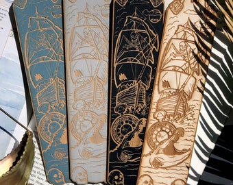 May include: Three wooden bookmarks with a pirate ship design. The bookmarks are in blue, black, and natural wood. Each bookmark features a skull and crossbones, a pirate ship, and a compass.