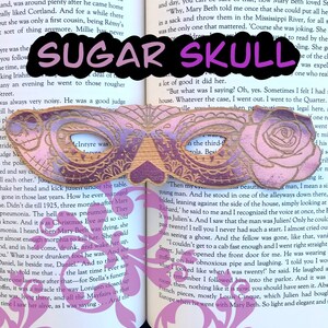 May include: A wooden sugar skull bookmark with a pink and purple gradient. The bookmark features intricate carvings of roses and other floral designs. The text "SUGAR SKULL" is printed in purple letters on the bookmark.