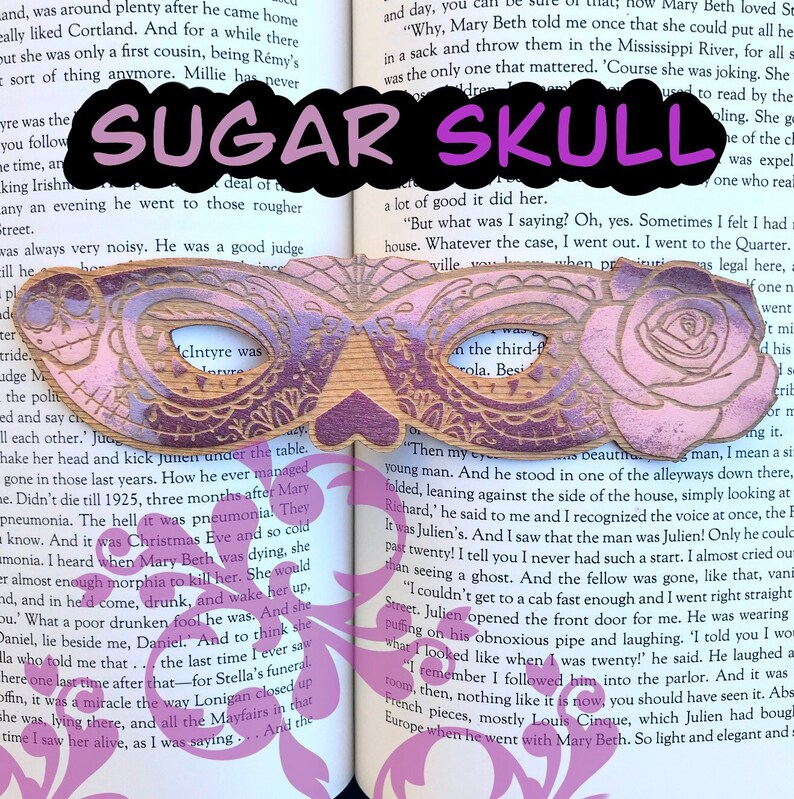 May include: A wooden sugar skull bookmark with a pink and purple gradient. The bookmark features intricate carvings of roses and other floral designs. The text "SUGAR SKULL" is printed in purple letters on the bookmark.