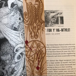 May include: A wooden bookmark with a carved image of a skull with tentacles. The bookmark is stained with red paint and has a pentagram symbol on the bottom. The text "FOR Y' HA-NTHLEI" and "OHN GLASBY" is visible on the page behind the bookmark.