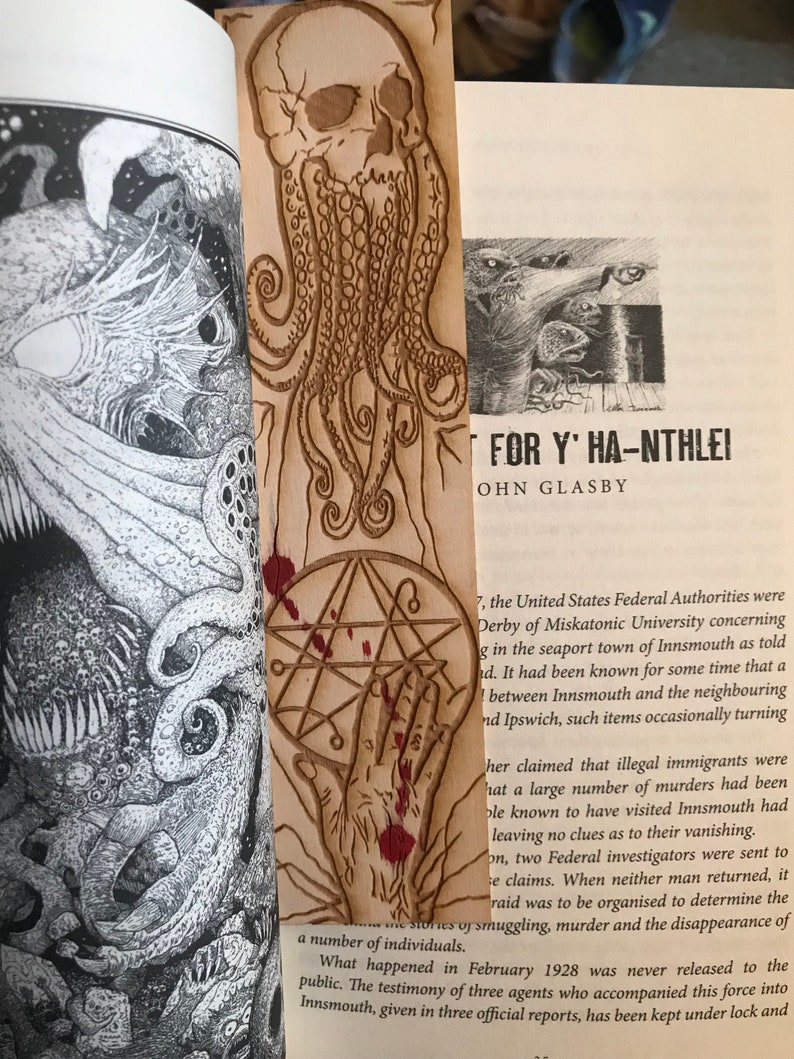 May include: A wooden bookmark with a carved image of a skull with tentacles. The bookmark is stained with red paint and has a pentagram symbol on the bottom. The text "FOR Y' HA-NTHLEI" and "OHN GLASBY" is visible on the page behind the bookmark.