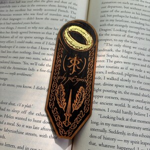 May include: A wooden bookmark with a black and gold design. The bookmark features a ring with an inscription that reads "The One Ring" and a sword with two leaves. The inscription "J.R.R. Tolkien" is also visible on the bookmark.