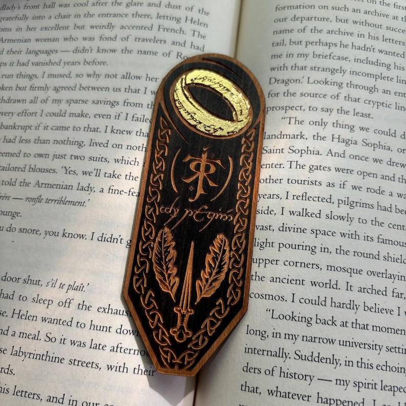 May include: A wooden bookmark with a black and gold design. The bookmark features a ring with an inscription that reads "The One Ring" and a sword with two leaves. The inscription "J.R.R. Tolkien" is also visible on the bookmark.