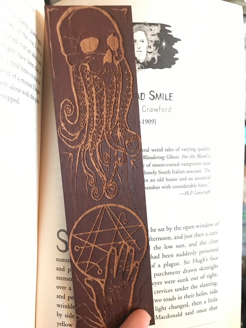 May include: A wooden bookmark with a dark brown finish. The bookmark features a detailed carving of a skull with tentacles and a pentagram. The text "AD SMILE Crawford -1909]" is visible on the bookmark.