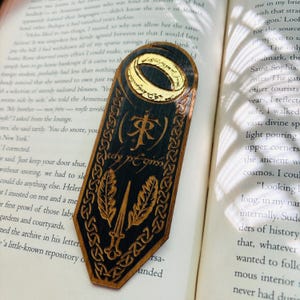 May include: A wooden bookmark with a dark blue background and gold accents. The bookmark features a depiction of the One Ring from the Lord of the Rings, along with the inscription "The Fellowship of the Ring".