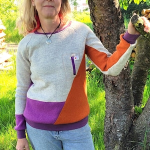Könnte beinhalten: Eine Frau in einem mehrfarbigen Pullover mit einer aufgesetzten Tasche steht vor einem Baum. Sie trägt eine blaue Jeans und Turnschuhe.