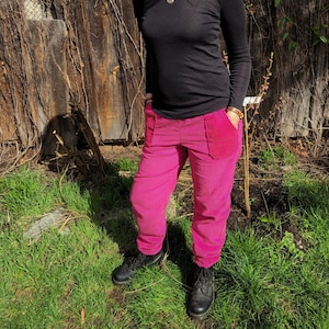 Könnte beinhalten: Eine Frau trägt ein schwarzes Langarmshirt und eine rosa Cordhose. Sie steht vor einer Holzwand mit Ranken.
