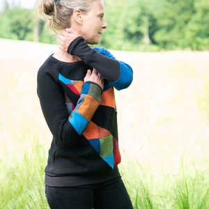 Könnte beinhalten: Eine Frau trägt einen schwarzen Pullover mit einem bunten Patchwork-Muster. Der Pullover hat ein blaues, oranges, grünes und rotes Muster. Sie steht auf einem Feld mit hohem Gras.