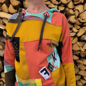Könnte beinhalten: Ein bunter Patchwork-Hoodie mit einem schwarzen Kordelzug. Der Hoodie besteht aus verschiedenen Stoffen und hat eine Vielzahl von Farben, darunter Gelb, Orange, Rot, Grün und Blau. Außerdem sind Patches auf den Hoodie genäht, darunter ein weißer Patch mit einem schwarzen Buchstaben "F" und ein blauer Patch mit einem weißen Buchstaben "K".