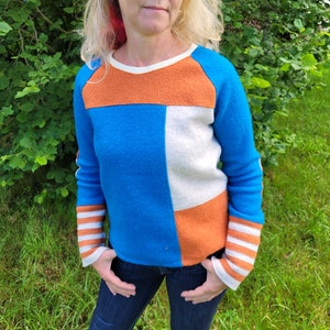 Könnte beinhalten: Ein blauer, orangefarbener und weißer Farbblockpullover mit gestreiften Ärmelbündchen. Der Pullover hat einen Rundhalsausschnitt und lange Ärmel.