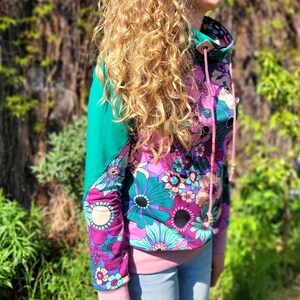 May include: A pink and green floral print sweatshirt with a high neck and a drawstring. The sweatshirt has a teal green panel on the shoulder and a pink drawstring.