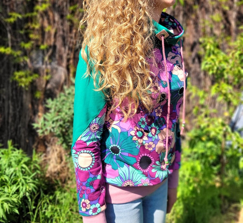 May include: A pink and green floral print sweatshirt with a high neck and a drawstring. The sweatshirt has a teal green panel on the shoulder and a pink drawstring.