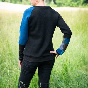 May include: A woman wearing a black jumper with blue and green patchwork on the sleeves and black jeans. She is standing in a field of long grass.