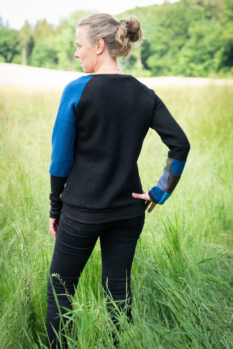 May include: A woman wearing a black jumper with blue and green patchwork on the sleeves and black jeans. She is standing in a field of long grass.