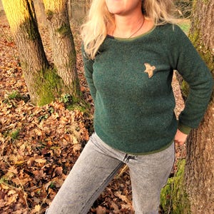 Könnte beinhalten: Ein dunkelgrüner Pullover mit einem braunen, vogelartigen Aufnäher auf der linken Brust. Der Pullover hat einen gerippten Ausschnitt und Bündchen. Die Person, die den Pullover trägt, steht in einer Waldumgebung.