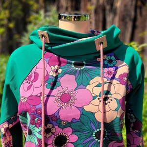 May include: A teal green hooded sweatshirt with a floral print featuring pink, purple, and white flowers. The hoodie has a drawstring with pink cords and metal eyelets.