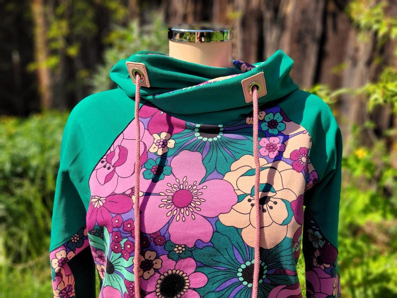 May include: A teal green hooded sweatshirt with a floral print featuring pink, purple, and white flowers. The hoodie has a drawstring with pink cords and metal eyelets.