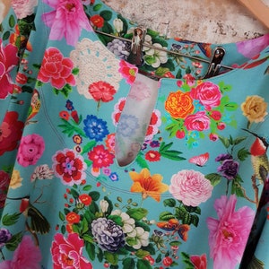 Könnte beinhalten: Eine türkisfarbene Bluse mit einem lebendigen Blumenmuster aus rosa, roten, orangen und gelben Blüten, grünen Blättern und kleinen Vögeln. Das Kleidungsstück hat einen Rundhalsausschnitt mit einem zentralen Schlüssellochausschnitt und cremefarbener Spitze.