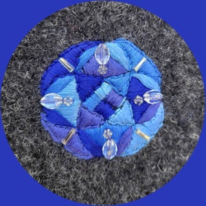 Könnte beinhalten: Ein blaues und violettes geometrisches Stickmuster mit klaren Kristallperlen auf einem grauen Filzgrund.