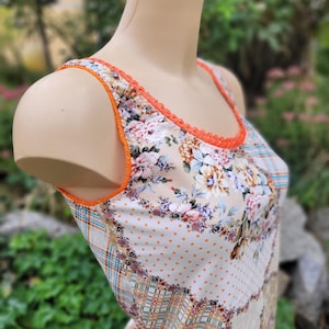Könnte beinhalten: Ein ärmelloses Tanktop mit Patchwork-Design, bestehend aus Blumenmustern, orangefarbenen Punkten und karierten Abschnitten. Der Ausschnitt und die Armausschnitte sind mit einer leuchtend orangefarbenen Bogenkante verziert, auf beigem Stoff.