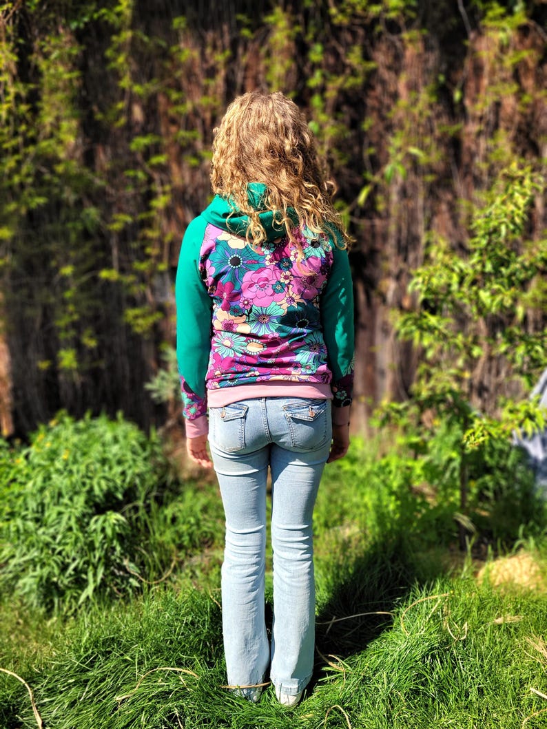 May include: A woman wearing a green and pink floral print hoodie with a hood and long sleeves. She is wearing blue jeans and standing in a grassy area.