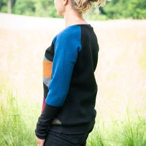 May include: A woman wearing a black and blue sweater with colourful patchwork details. She is standing in a field of tall grass.