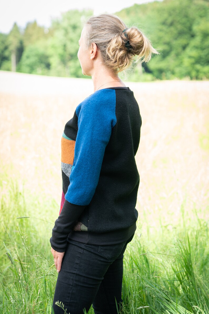 May include: A woman wearing a black and blue sweater with colourful patchwork details. She is standing in a field of tall grass.