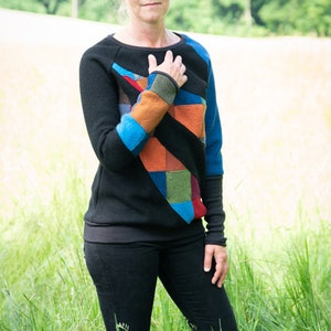 May include: A woman wearing a black jumper with a colourful patchwork design. The jumper has a geometric pattern with blue, orange, green, and red accents. She is standing in a field of tall grass.