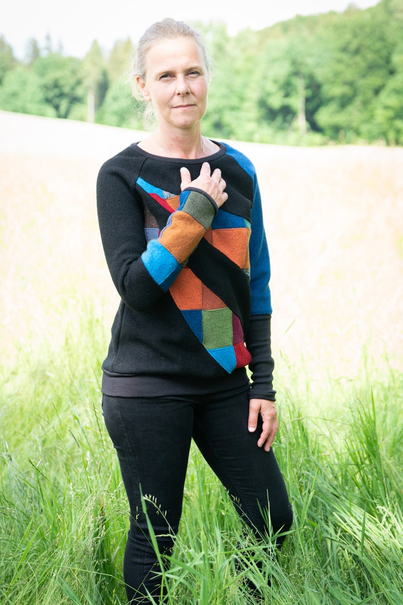 May include: A woman wearing a black jumper with a colourful patchwork design. The jumper has a geometric pattern with blue, orange, green, and red accents. She is standing in a field of tall grass.