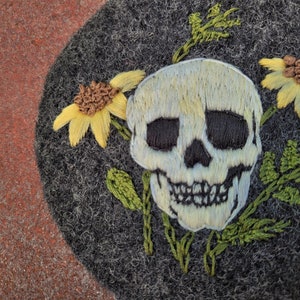Könnte beinhalten: Dunkelgrauer Filzbarett mit einem gestickten Totenkopf-Design. Der Totenkopf ist weiß mit schwarzen Augenhöhlen und Nase, umgeben von gelben Sonnenblumen und grünen Blättern. Ein einzigartiges, handgefertigtes Accessoire.