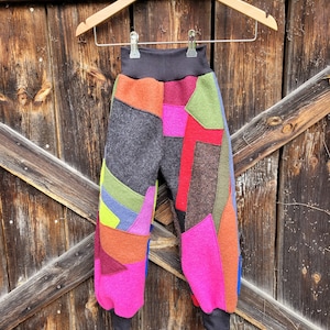 Könnte beinhalten: Patchwork-Hose mit verschiedenen Farben und Mustern, darunter Pink, Rot, Grün, Braun und Grau. Die Hose hängt an einem Holzbügel.