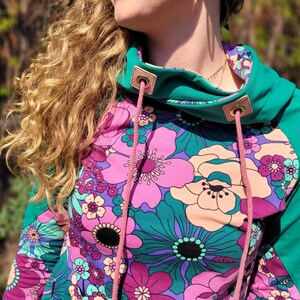 May include: A teal green and pink floral print hoodie with a pink drawstring. The hoodie has a high neck and a relaxed fit.