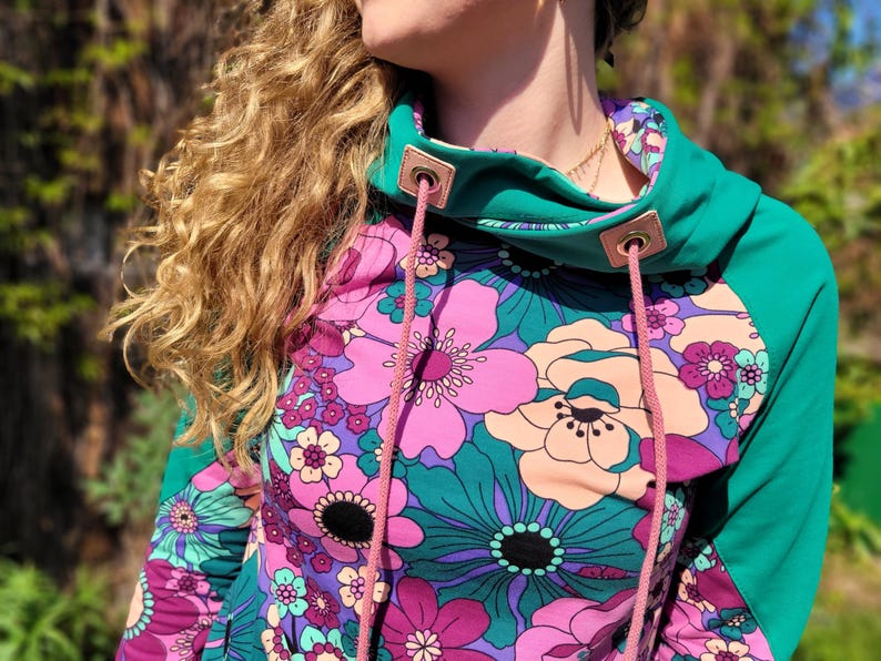 May include: A teal green and pink floral print hoodie with a pink drawstring. The hoodie has a high neck and a relaxed fit.