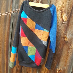 May include: A black sweatshirt with a colourful patchwork design on the front. The patchwork is made up of squares and rectangles in various shades of blue, red, orange, green, and brown. The sweatshirt has a blue long sleeve and a black long sleeve.