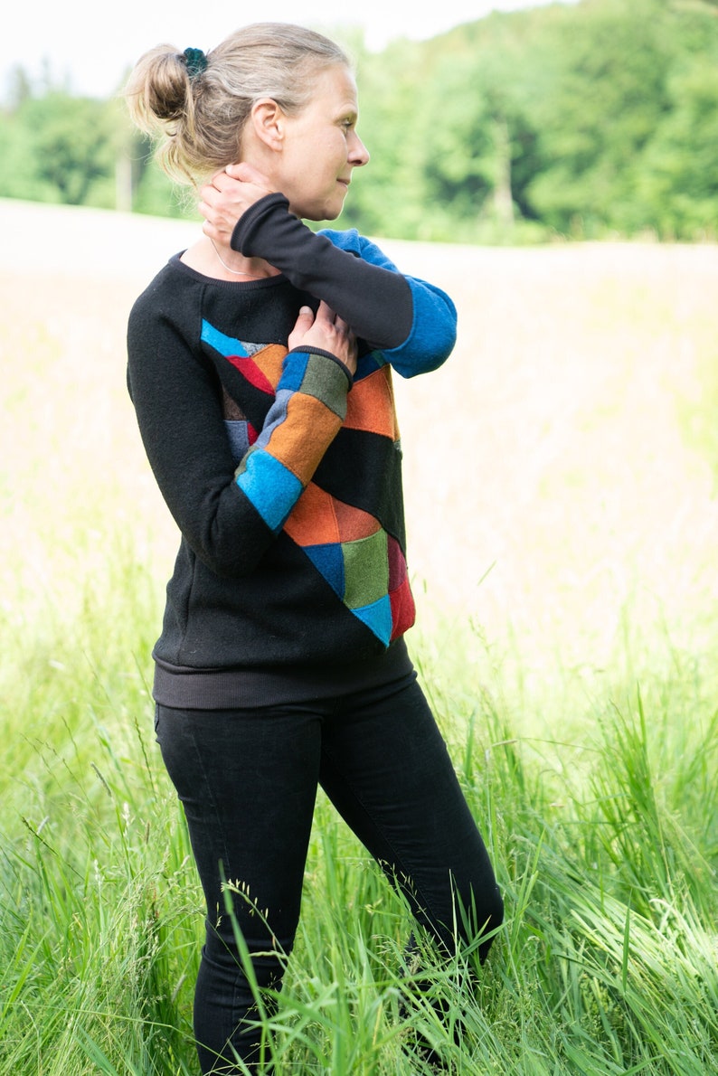 May include: A woman wearing a black jumper with a colourful patchwork design. The jumper has a blue, orange, green and red pattern. She is standing in a field of long grass.