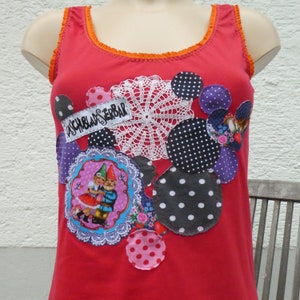 Könnte beinhalten: Rotes Tanktop mit verschiedenen bunten Stoffapplikationen, darunter Punkte, eine gehäkelte Spitze und eine Szene mit zwei Gnomen. Der Text "unschlagbar" ist auf der Applikation sichtbar.