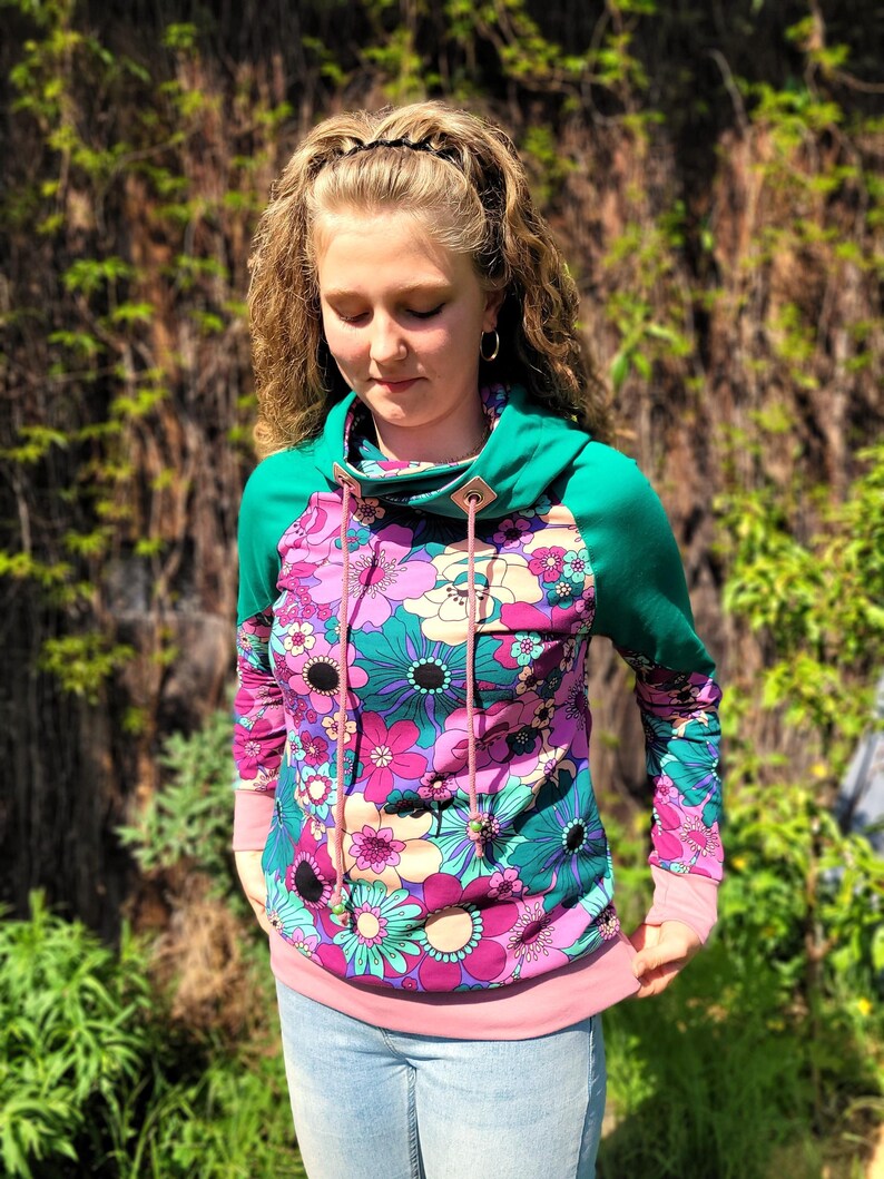 May include: A teal and pink long-sleeved sweatshirt with a floral print and a cowl neck. The sweatshirt has a drawstring with pink cord and a small button on the neck.