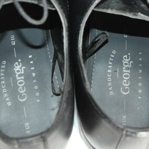 May include: Close-up of the inside of two black leather shoes. The shoes are labelled "HANDCRAFTED - George. FOOTWEAR" and have size markings in UK and EU sizes.
