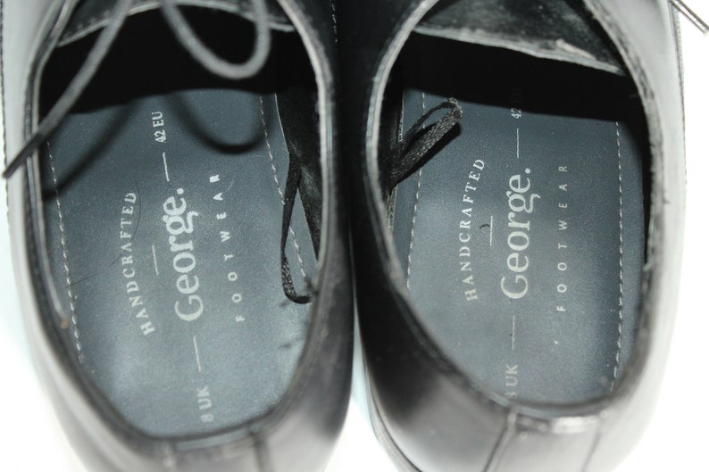 May include: Close-up of the inside of two black leather shoes. The shoes are labelled "HANDCRAFTED - George. FOOTWEAR" and have size markings in UK and EU sizes.