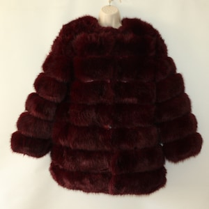 Może przedstawiać: Bordowy płaszcz z imitacji futra, prezentowany na manekinie. Płaszcz ma warstwową, poziomą konstrukcję i długie rękawy. Pluszowy materiał ma bogaty, głęboki kolor, a płaszcz wygląda na miękki i ciepły. Odzież jest przeznaczona na zimną pogodę.