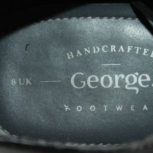 May include: Close-up of the inside of a black leather shoe. The insole is grey and has the text "HANDCRAFTED George. FOOTWEAR" and "8 UK" printed on it.