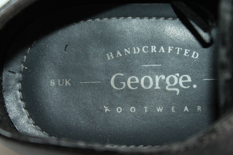 May include: Close-up of the inside of a black leather shoe. The insole is grey and has the text "HANDCRAFTED George. FOOTWEAR" and "8 UK" printed on it.
