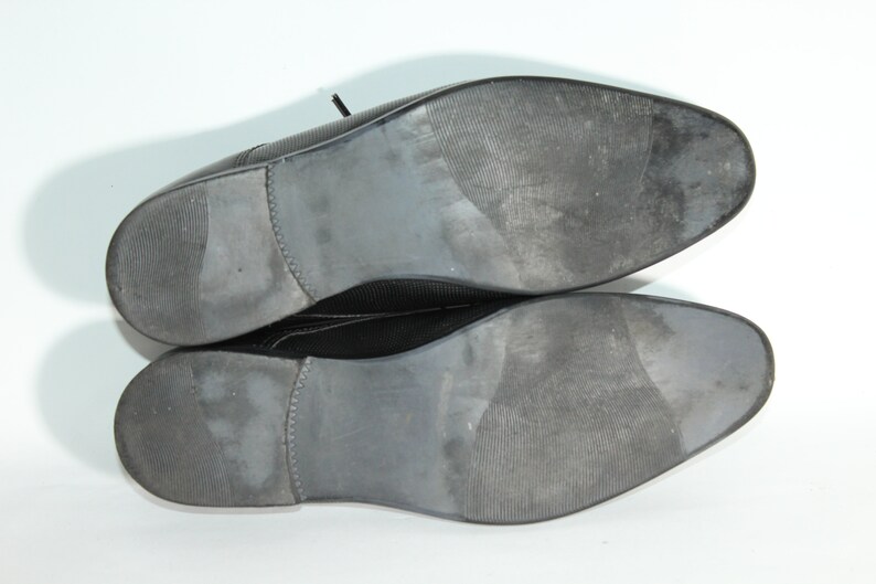 May include: A pair of black leather dress shoes with a textured sole. The shoes are shown from the bottom, with the soles facing the camera.