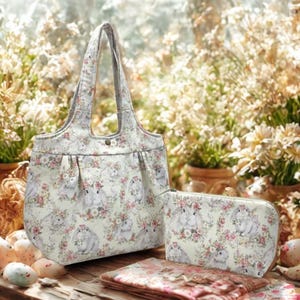 May include: A tote bag, clutch, and folded fabric featuring a floral and bunny print. The tote bag has a button closure and handles. The clutch has a zipper closure. The print includes bunnies wearing floral crowns and pink and white flowers.