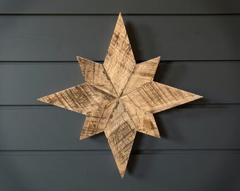 May include: A wooden star decoration with eight points, mounted on a dark blue-grey wall. The star is made of light brown, weathered wood. The wood grain is visible, adding texture to the star. The star's dimensions are approximately 60 cm.