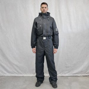 May include: Black one-piece snowsuit with a hooded collar, multiple pockets, and a belt. The snowsuit has a zipper closure and a small patch on the upper arm. The outfit is made of a durable, water-resistant material, perfect for winter sports and outdoor activities.