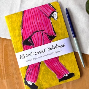 Peut inclure: Cahier A5 à couverture souple avec un fond jaune et une illustration rose d'une personne en costume. Le cahier contient 52 pages vierges et est illustré par Majuk. Un stylo bleu est posé à côté du cahier sur une surface en bois.