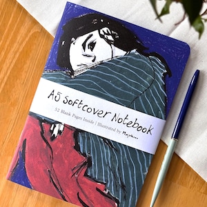 Peut inclure: Un carnet A5 à couverture souple avec une illustration d'une personne sur fond bleu. Le carnet est entouré d'une bande blanche avec le texte "A5 softcover Notebook". Un stylo bleu et argenté est posé à côté du carnet.