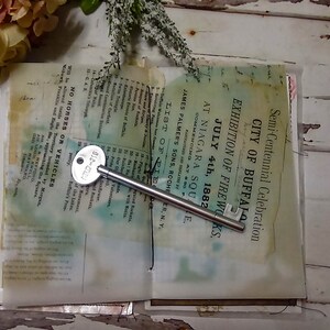 May include: An open book with a silver key on top of it. The book is old and has a faded, yellowed appearance. The text on the book reads "Semi-Centennial Celebration City of Buffalo Exhibition of Fireworks July 4th, 1882 at Niagara Square."