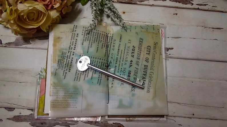 May include: An open book with a silver key on top of it. The book is old and has a faded, yellowed appearance. The text on the book reads "Semi-Centennial Celebration City of Buffalo Exhibition of Fireworks July 4th, 1882 at Niagara Square."